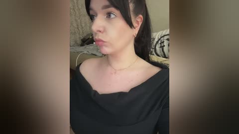 A video of a young woman with fair skin, dark hair, and an off-the-shoulder black top, wearing a dainty necklace. She gazes thoughtfully into the distance, set against a blurred background.