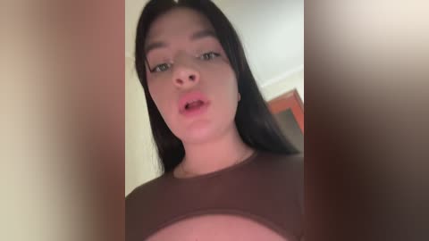 A video of a young woman with fair skin, straight black hair, and a round face. She wears a brown top and has a surprised expression. The background is blurred, with a hint of a wooden door.