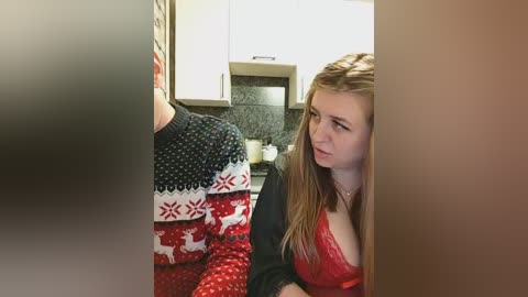 A video of a young woman in a red lace bra and black cardigan, looking through a fogged-up window in a modern kitchen.