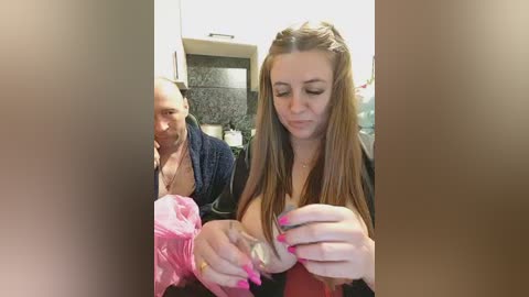 A video shows a young woman with long, straight brown hair and pink nails, holding a baby in a pink blanket. She wears a black top. The background features a kitchen with dark countertops and white cabinets.