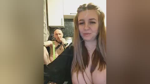 Video of a young, fair-skinned woman with straight, light brown hair and a black top, standing in a doorway. A man with a bald head, wearing a black shirt, sits in a kitchen with a dark countertop, appearing surprised.