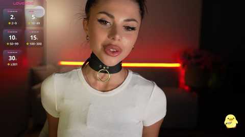Video of a young woman with light skin, dark hair in a bun, wearing a white T-shirt and black leather collar with a metal ring, set in a dimly-lit room with a red neon strip light in the background.