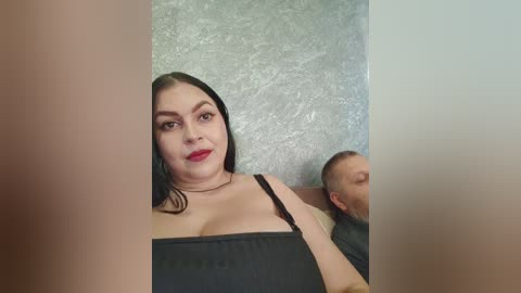 A video of a smiling woman with fair skin and long black hair in a black dress, standing beside a bald man in a dark shirt, both looking at a mirror.