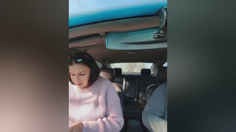 A video of a young woman with long black hair, wearing a light pink sweater, sitting in the back seat of a car, looking down at her phone.