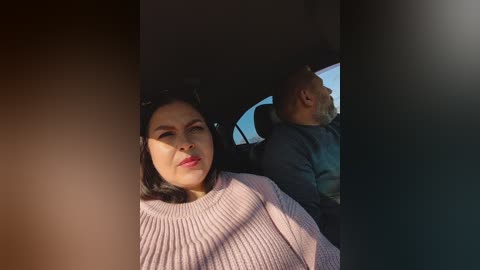 Video of a Latina woman with medium skin tone, short black hair, and red lipstick, wearing a pink knitted sweater, sitting in a car next to a bald, older Black man in a dark shirt.
