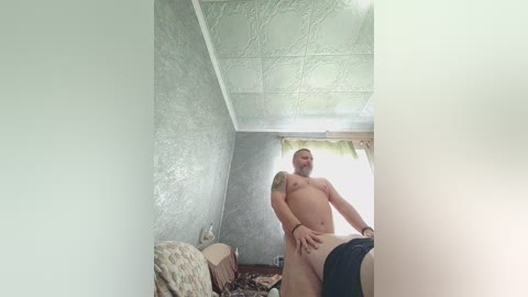 Video of a muscular, naked, bearded man with a tattoo, standing in a dimly lit, rustic room with textured walls, a bed with a patterned blanket, and a window with a sheer curtain.