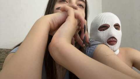 Video of a young woman with long dark hair, wearing a white balaclava, holding her bare foot close to her face, smiling. She has fair skin and is dressed in a casual, light-colored top. Background is a plain, light-colored wall.