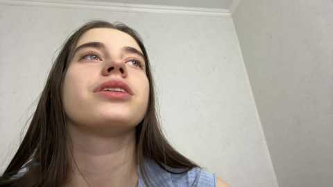 Video of a young woman with long, straight brown hair, fair skin, and blue eyes, wearing a light blue top, looking upward against a plain white wall.