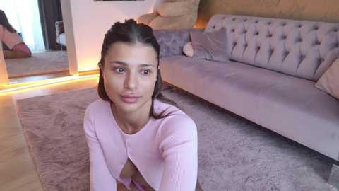 Video of a young woman with dark hair, wearing a pink cardigan, sitting on a plush rug in a modern living room with beige furniture.