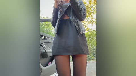 Video of a slender, light-skinned woman in a black leather jacket and mini-skirt, standing outside next to a car in daylight.