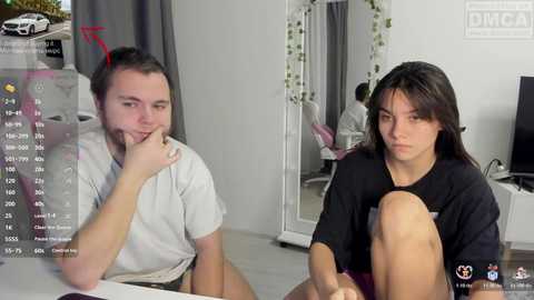 A video of a man and woman, both in casual clothes, sitting on a bed in a modern bedroom with white walls, plants, and a mirror.