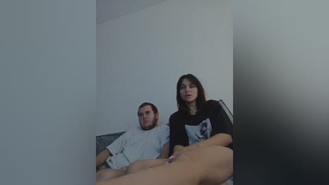 A video shows a man and woman, both casually dressed, sitting on a bed in a dimly lit, sparsely decorated room with white walls. The woman has long dark hair and wears a black t-shirt, while the man has short hair and a beard, wearing a white shirt.