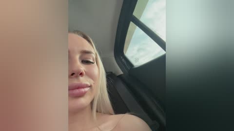 A video captures a close-up of a blonde woman with fair skin, slightly parted lips, and closed eyes, seated in a car, illuminated by daylight through the window.