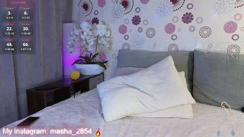 Video of a cozy bedroom with a gray bed, white pillows, floral-patterned wallpaper, white orchid, and a purple lamp on a wooden nightstand. Text overlay reads \"My Instagram mashup 2585.\