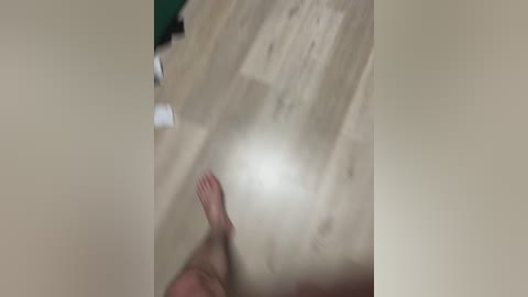 A blurry video of a wooden floor with a green object partially visible in the background. The image is slightly out of focus, with a hand and bare foot at the bottom center.