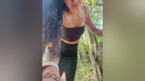 Video of a slender, dark-haired woman in a black sports bra and leggings, hiking outdoors in a forest with sunlight filtering through the trees.