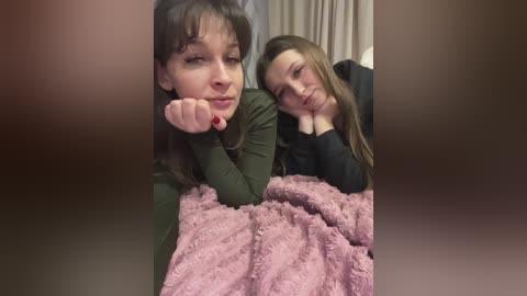 Video of two young women with fair skin, one with long brown hair, the other with long blonde hair, lying on a bed, wrapped in a pink blanket, with a cozy, indoor setting.
