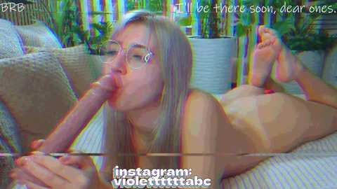 A video captures a nude blonde woman with glasses performing oral sex on a large, erect penis. She is on a beige couch, with a blurred green background. Text overlays say \"I'll be there soon, dear ones\" and \"instagram: violertrttabc.\