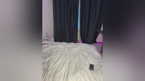 A video of a dimly lit bedroom with a messy bed covered in white sheets, black curtains, and a partially visible desk in the background.