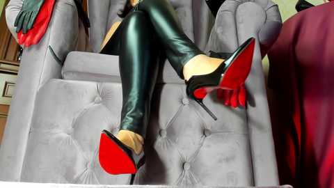 Video of a person in black leather leggings and red stiletto heels, seated on a plush grey armchair, with red gloves and a red scarf visible.