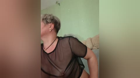 A video of a light-skinned woman with short, gray hair, wearing a sheer black top and a black necklace, leaning against a beige couch in a green-painted room.