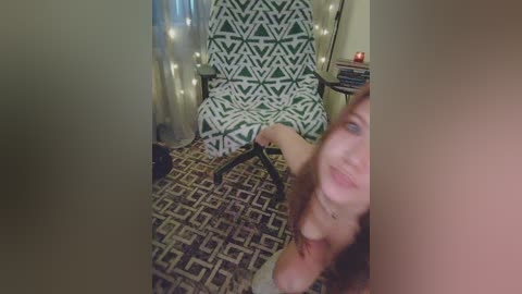 A video of a woman with fair skin and red hair, sitting on a patterned green and white chair, wearing a white top, in a dimly lit room with patterned carpet, candles, and a wooden side table.