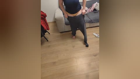 A video of a person in a dark shirt and gray sweatpants, walking barefoot in a living room with a beige couch, wooden floor, and a red jacket draped over a chair.