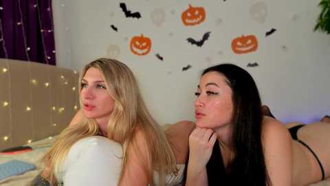 Video of two women with long hair, one blonde, one brunette, lying on a bed, wearing lingerie, in a Halloween-themed room with carved pumpkins, bats, and string lights.