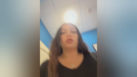 Video of a woman with long dark hair, wearing a black top, standing in a room with blue walls and a white ceiling. The image is slightly blurred and has a soft focus.