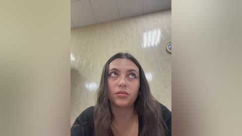 Video of a woman with long dark hair and light skin, wearing a black shirt, standing in a tiled bathroom with beige walls and a round clock on the ceiling.