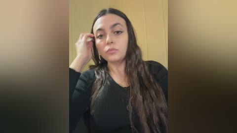 Video of a young woman with long, dark hair, wearing a black sweater, against a blurred wooden background. She has a neutral expression, and her right hand touches her hair.