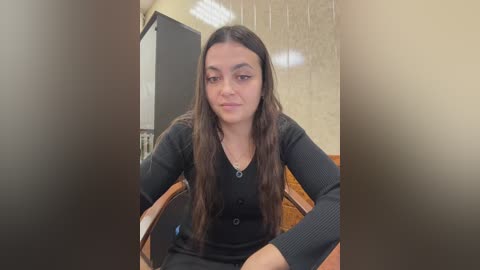 Video of a young woman with long dark hair, wearing a black sweater, sitting in a wooden chair in a beige-tiled room with fluorescent lighting.