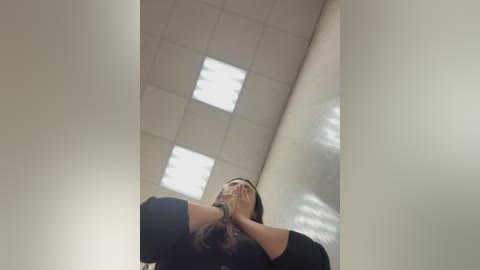 Video of a woman with long, dark hair, wearing a black top, standing in a dimly lit, white-tiled hallway with fluorescent lights overhead, hands covering her face.