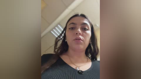 Video of a young woman with light skin and long dark hair, wearing a black ribbed sweater and a necklace. She stands in a dimly lit, beige-colored room with a door and window visible.