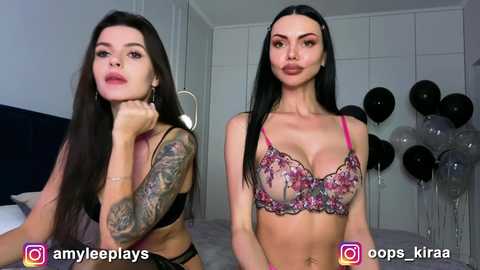 Video of two women with long dark hair, one with tattoos, in lingerie, standing in a modern bedroom with white walls and black balloons.
