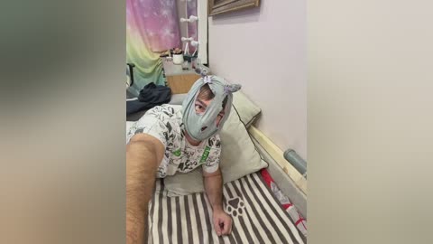 A video captures a person with a grey mask, lying on a striped bed in a colorful, cluttered room with pastel walls, a rainbow tapestry, and a furry toy.
