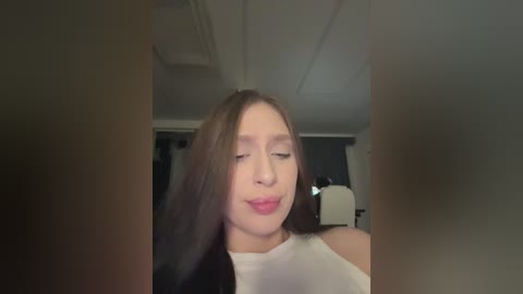 Video of a young woman with long brown hair, fair skin, and closed eyes, wearing a white sleeveless top, in a dimly lit room with a ceiling fan.