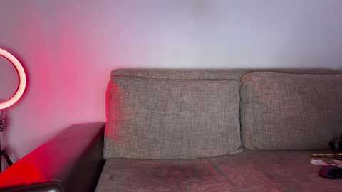 Video of a beige fabric sofa against a plain white wall, illuminated by a red light from a round ring light on the left, with a small, partially visible object on the right.