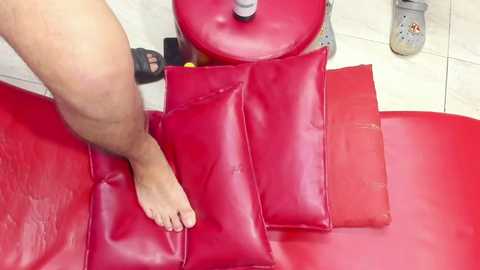A video of a person's foot stepping on a vibrant red leather couch. The couch features two matching red cushions. In the background, there's a ceramic mug and a bottle. The scene is indoors on a tiled floor.