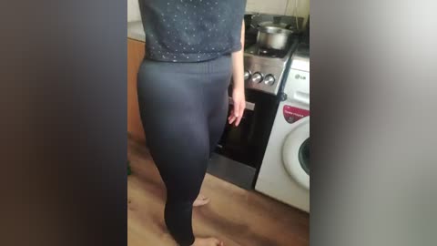 Video of a person with fair skin wearing a dark, polka-dotted top and tight, black leggings, standing in a kitchen with a white washer, wooden floor, and stove.