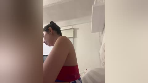 A video of a woman in a red strapless top, sitting on a bed, with a white wall and a beige ceiling in the background.