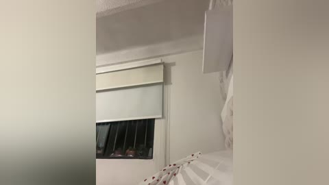 Video of a bedroom with a white ceiling, beige walls, and a partially open window with a white shade. A white bed with a red patterned sheet is visible on the right side.