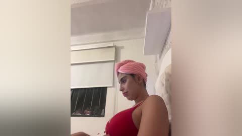 Video of a young woman with light brown skin, wearing a red tank top and pink towel turban, leaning against a white wall in a dimly lit room with a window and blinds.