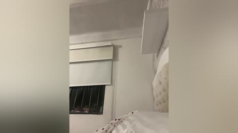 Video of a minimalist bedroom with a white bed, a beige tufted headboard, and a closed white window blind. The walls are plain white, and the room has a cozy, understated feel.