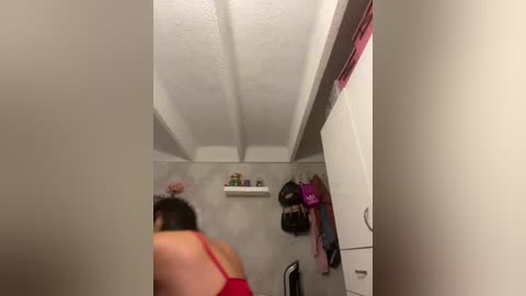 A video of a small, cramped room with a woman in a red top, sitting on a chair. White walls, a shelf with toiletries, and a hanging bag with pink items.