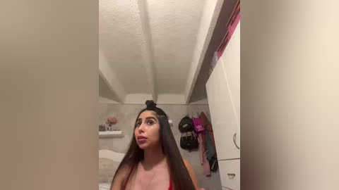 A video of a young woman with long brown hair, wearing a red top, standing in a small, sparsely furnished bedroom with white walls, exposed ceiling beams, and a pink jacket hanging on the closet door.