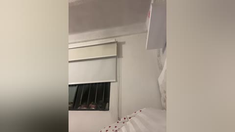 Video of a simple bedroom with a white bed, beige window blind, and white wall. The room is sparsely furnished, with minimal decor, suggesting a clean and functional space.