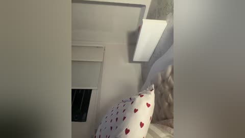 Video of a cozy attic bedroom with a white quilted headboard, a white pillow with red heart patterns, a small window with a closed blind, and a modern, rectangular wall light.