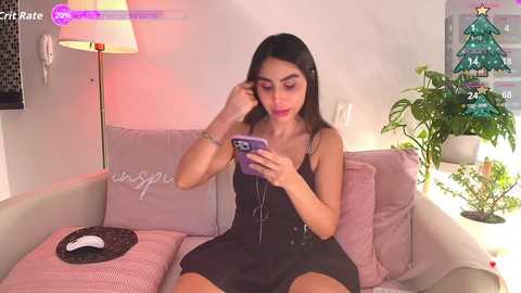 A video of a Latina woman with long black hair, wearing a black dress, using a smartphone while sitting on a pastel couch with a green plant in the background.