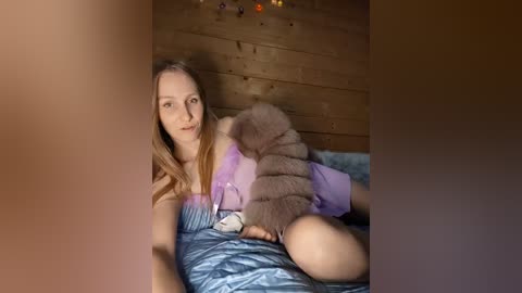 A video of a young woman with long, straight hair lying on a bed, wearing a lavender shirt. She holds a large, fluffy brown bear. The background features wooden walls and string lights.